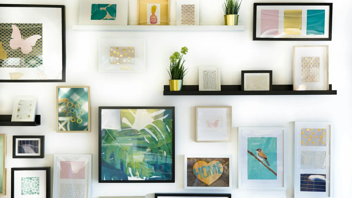 A sunlit white wall arranged with many small framed prints, pastel artworks and potted plants on slim shelves.
